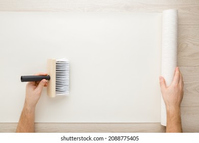 Young Adult Man Hand Using Brush And Applying Glue On Wallpaper Sheet On Wooden Table Background. Closeup. Repair Work Of Home. Point Of View Shot. 