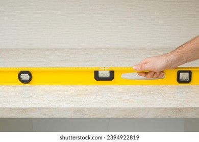 Young adult man hand using yellow spirit level and measuring horizontal surface of beige stone tabletop. Assembling new kitchen furniture. Closeup. Front view. Renovation process. - Shutterstock ID 2394922819