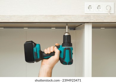 Young adult man hand using professional battery screwdriver and screwing screw in metal base cabinet frame and tabletop. Assembling new kitchen wooden furniture. Closeup. Front view. - Shutterstock ID 2394923471