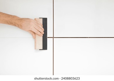 Young adult man hand using rubber trowel and grouting seams with paste between white ceramic tiles on wall or floor. Closeup. Renovation process. Repair work of home.