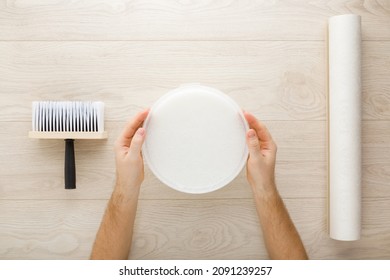 Young Adult Man Hands Holding Bucket With Glue. Roll Of Wall Paper, Brush On Light Wooden Table Background. Closeup. Point Of View Shot. Preparing For Repair Work Of Home. Ready To Glue Wallpaper. 