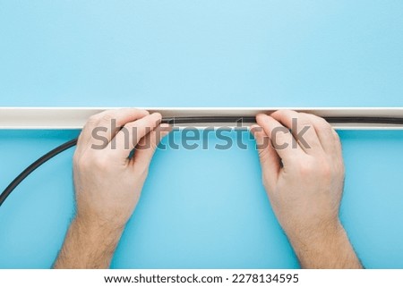 Young adult man hands putting dark black new wire in white plastic channel on light blue wall background. Pastel color. Closeup. Point of view shot. Electrician working.