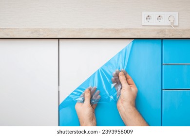 Young adult man hands removing blue protective polythene film from new kitchen cabinet doors under table top. Closeup. Front view. - Shutterstock ID 2393958009