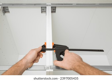 Young adult man hands using f speed clamp and pressing white boards of base cabinet frame. Assembling new kitchen wooden furniture. Renovation process. Closeup. Point of view shot.