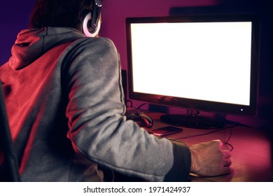 Young Adult Man With Headset Sitting At Desk Gaming Setup Playing Video Games Illuminated By Red And Purple Lights. Monitor With Empty Blank Screen For Designs.