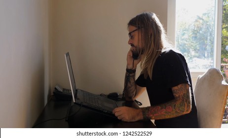 Young adult man with long blonde hair and a beard working from home on a laptop at a desk in his apartment answering a phone call on his home landline cord phone nodding and hanging up with glasses on