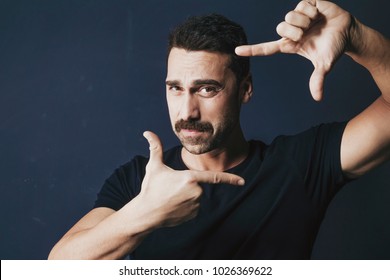 Young Adult Man With Moustache Framing His Face With His Hands. Handsome Bearded Man Wearing A Black T-shirt, Isolated On Dark Background. Close Up.
