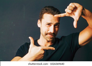 Young Adult Man With Moustache Framing His Face With His Hands. Handsome Bearded Man Wearing A Black T-shirt, Isolated On Dark Background. Close Up.
