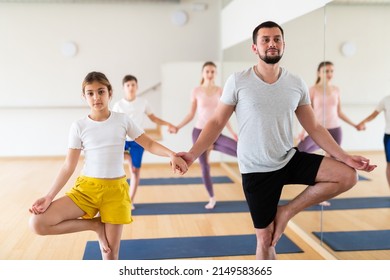 Young Adult Man Practicing Yoga In Pair With Teen Daughter, Performing Double Tree Pose During Family Workout At Gym