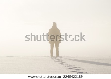 Young adult man silhouette standing in nature mist and looking far away. Thinking about life. Back view. Fresh human boots footprints in fresh deep snow. Cold snowy winter day.