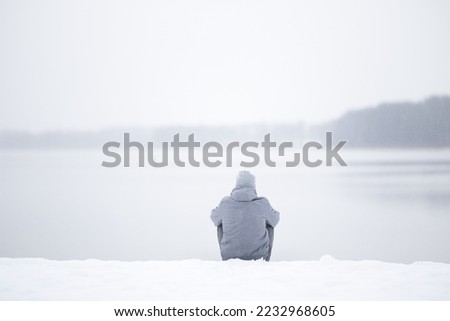Young adult man sitting alone on snow at lake shore and looking far away. White cold snowy winter day. Thinking about life. Peaceful atmosphere in nature. Back view.