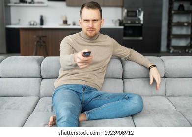 Young Adult Man Is Sitting On Sofa With A TV Remote In The Hand, Trying To Watch Something On TV, Saw Himself In The Program, Spending Time At Home, Tuning The TV, Fast Forward Or Rewind, Foreground