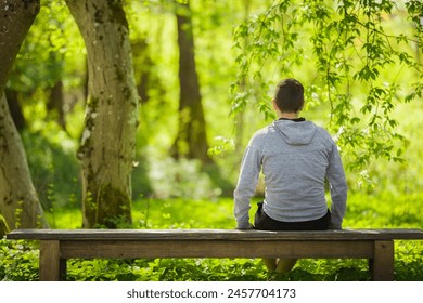 Young adult man sitting on bench and staring at green leaves, grass and trees. Thinking about life at beautiful park in sunny spring day. Spending time alone in nature. Peaceful atmosphere. Back view. - Powered by Shutterstock