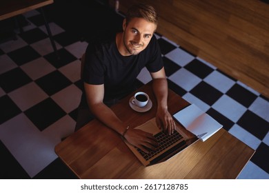 Young adult man sitting at wooden café table, typing on laptop with coffee cup and notebook. Freelancer, productivity, remote work, workspace, interior, overhead, candid