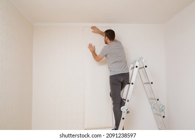 Young Adult Man Standing On Metal Ladder And Applying New Wallpaper On White Wall In Room. Repair Work Of Home. Making Interior Change. Back View.