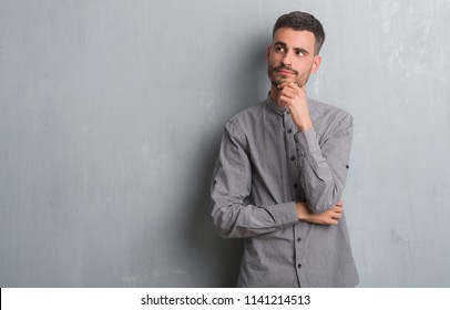 Young adult man standing over grey grunge wall with hand on chin thinking about question, pensive expression. Smiling with thoughtful face. Doubt concept. - Shutterstock ID 1141214513