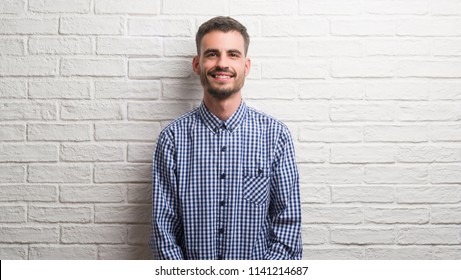 Young adult man standing over white brick wall with a happy face standing and smiling with a confident smile showing teeth