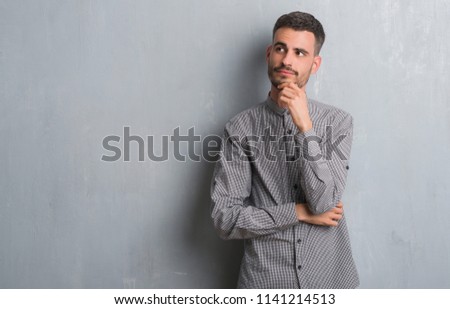 Young adult man standing over grey grunge wall with hand on chin thinking about question, pensive expression. Smiling with thoughtful face. Doubt concept.