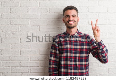 Young adult man standing over white brick wall showing and pointing up with fingers number two while smiling confident and happy.