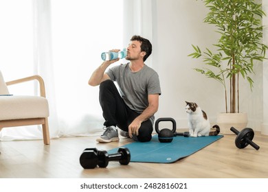 Young adult man training with weights in the living room, resting to hydrate himself by drinking water or isotonic drink with his pet cat, house with minimalist decoration - Powered by Shutterstock