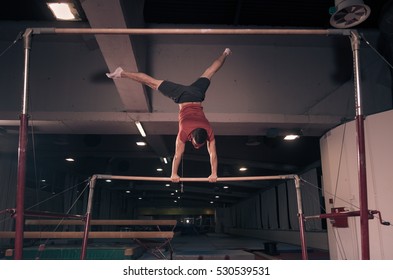 Young Adult Man, Two Hand Cartwheel. Gymnast Holding Hands, Horizontal Bar, Legs In Air. Indoors Dark Hall, Gymnastics Equipment.