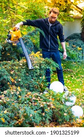 Young Adult Man Using A Cordless, Electric Leaf Blower In A Garden. Garden Works. Autumn, Fall Gardening Works In A Backyard, On A Lawn, A Grass, Bushes.