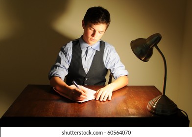 A Young Adult Man Writing A Letter On A Desk With A Lamp.