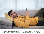 Young adult mature man using smart phone resting lying down on sofa couch at home messaging on mobile phone Guy with eyeglasses relaxing at home surfing social media buying purchasing paying ordering