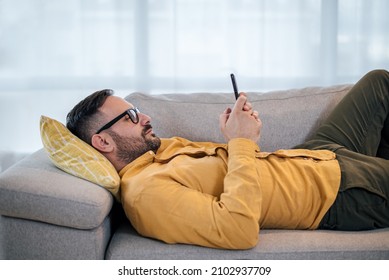 Young adult mature man using smart phone resting lying down on sofa couch at home messaging on mobile phone Guy with eyeglasses relaxing at home surfing social media buying purchasing paying ordering