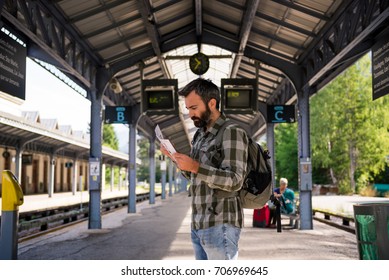 Young adult men commuter waiting for train in mountain train station reading timetable in sunny summer day.