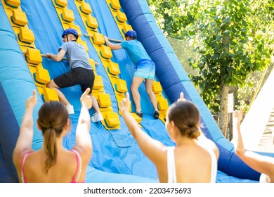 Young And Adult Men Using Sticks To Climb Up On Slide In Adventure Park. Women Supporting Them, Raising Hands And Gesturing.