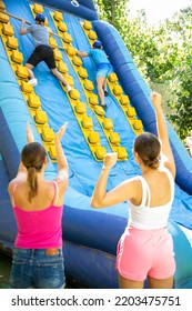 Young And Adult Men Using Sticks To Climb Up On Slide In Adventure Park. Women Supporting Them, Raising Hands And Gesturing.