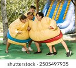 Young and adult men and women in large sumo suits pushing each other, having fun in adventure park.