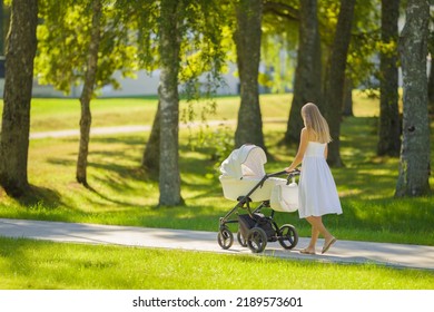 Young adult mother in dress pushing white baby stroller and walking on sidewalk at town park in warm sunny summer day. Spending time with infant and breathing fresh air. Enjoying stroll.