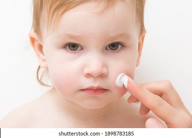 Young Adult Mother Finger Applying White Moisturizing Cream On Toddler Cheek. Daily Care About Baby Face Skin. Closeup. Front View. Isolated On Light Gray Background.