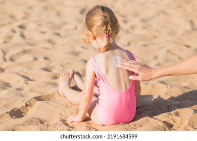 Young Adult Mother Hand Applying White Sunscreen Lotion On Little Girl Back. Child Sitting On Sand. Skin Protection. Safety Sunbathing In Hot Sunny Day At Beach. Rear View. Closeup.