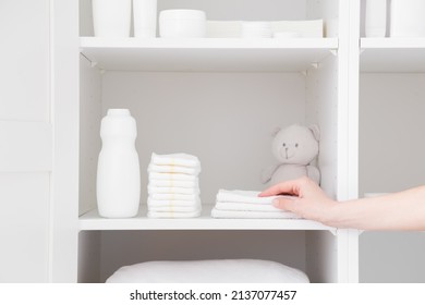 Young Adult Mother Hand Taking White Towel From Inside Wardrobe. Bottle Of Talcum Powder, Stack Of New Nappies And Teddy Bear On Shelf. Preparing For Baby Diaper Changing. Hygiene Goods. Closeup.