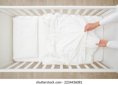 Young adult mother hands changing white blanket cover in baby crib. Regular bed linen change. Closeup. Top view. 