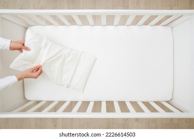 Young adult mother hands changing white cotton cover on pillow in baby crib. Regular bed linen change. Closeup. Top view.