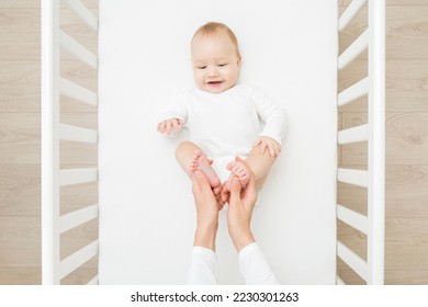 Young adult mother hands holding and massaging infant cute small bare feet in white crib at home room. Lovely emotional moment. Closeup. Parent playing with happy adorable smiling baby boy. Top view.