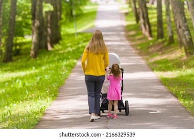 Young Adult Mother And Little Daughter Pushing White Baby Stroller And Walking At Town Park In Warm Sunny Summer Day. Spending Time Together And Breathing Fresh Air. Enjoying Stroll. Two Child Mom.