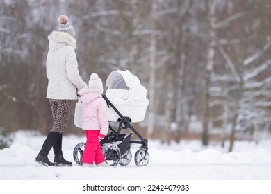 Young adult mother and little daughter pushing white baby stroller and walking on snow covered sidewalk at park in cold winter day. Spending time together. Enjoying stroll. Two child mom. Back view.
