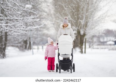 Young adult mother and little daughter pushing white baby stroller and walking on snow covered sidewalk at park in cold winter day. Spending time together. Enjoying stroll. Two child mom. Front view.