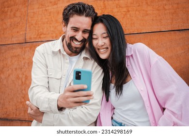 Young adult multiracial couple having fun and laughing using a smartphone outside. Cheerful pair of friends smiling and watching funny videos on a cellphone app. Two happy people using a mobile phone - Powered by Shutterstock