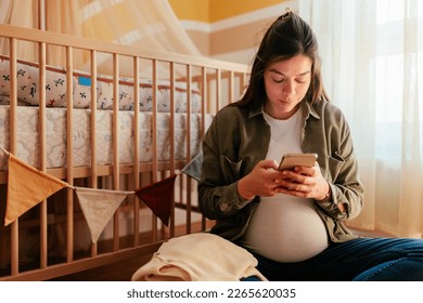 A young adult pregnant woman is at home using her cellphone while sitting in the nursery room. - Powered by Shutterstock