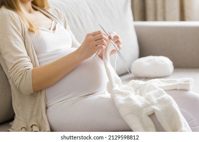 Young Adult Pregnant Woman Sitting On Sofa At Living Room And Knitting First Warm Bodysuit For Newborn From White Wool. Making Clothes For Future Baby In Pregnancy Time. Side View.