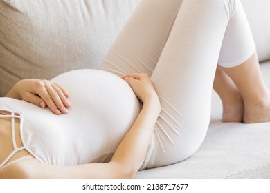 Young adult pregnant woman in white clothes lying on back and touching big belly and feeling her baby move. Relaxing on sofa. Baby expectation. Pregnancy time. Closeup.  - Powered by Shutterstock
