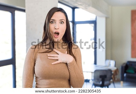 young adult pretty woman feeling shocked, astonished and surprised, with hand on chest and open mouth, saying who, me?