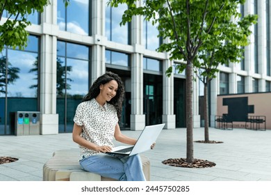 A young adult is seated outdoors, working intently on a laptop in a casual setting outside a contemporary office building, surrounded by nature. - Powered by Shutterstock