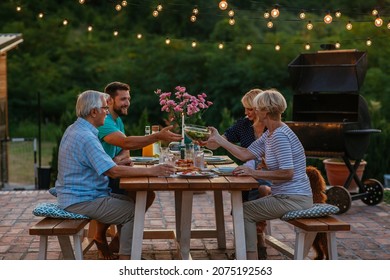 Young Adult And Senior Couple Sitting At The Dining Table In The Backyard And Enjoying Dinner